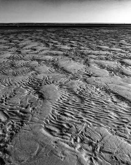 A high-contrast monochrome photo of wet sand ripples at a beach. Detailed ridges and grooves cut through the wet sand and the beach extends toward a distant horizon.