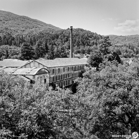 A black and white photograph depicts an old Textile Mill nestled within a dense, hilly landscape. A tall chimney rises from the building's roofline, contrasting with the surrounding greenery and a glimpse of another structure partially obscured by foliage. The afternoon light illuminates the scene, creating a sense of quiet history and natural beauty.