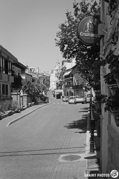 A street lined with buildings, featuring a sign for Cruzcampo beer. The scene is captured in black and white, showcasing a clear sky above, with trees providing shade along the path. A few vehicles are parked on the side of the road.