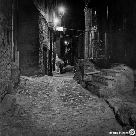 A dimly lit, narrow street winds through the historic village of Miranda del Castañar, its stone walls rising on either side. The rough cobblestone pavement leads into the darkness, illuminated by a single street lamp at the far end. The scene evokes a sense of quiet solitude and old-world charm.