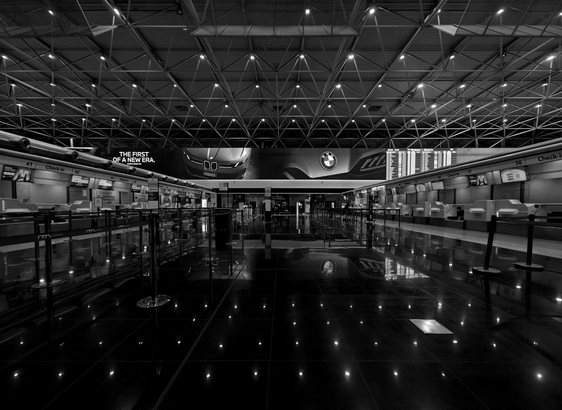 A photo of an airport check-in hall with a reflective black tile floor and a geometric metal structure and small bright lights in the ceiling. The hall is mostly empty, looking deserted.