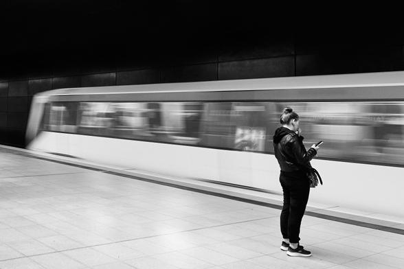 Eine junge Frau steht auf einem Bahnsteig und blickt auf ihr Handy. Direkt vor ihr fährt eine U-Bahn ins Bild. Durch eine längere Belichtungszeit ist die Bahn verschwommen dargestellt.