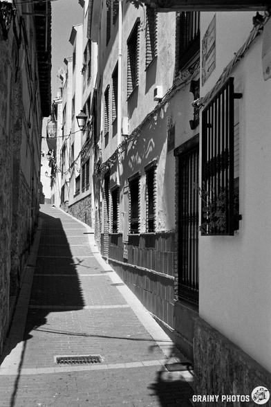 A narrow uphill alleyway lined with traditional, whitewashed buildings, featuring barred windows and cobblestone pavement, illuminated by sunlight casting shadows.