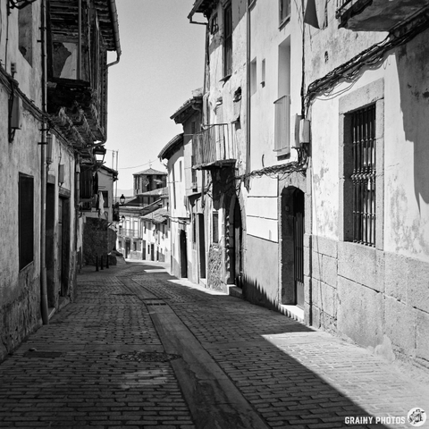 A black and white photograph captures a narrow, cobbled street lined with old stone buildings in Béjar, Spain. The afternoon light casts long shadows down the street, highlighting the textures of the buildings and pavement, creating a sense of quiet history.