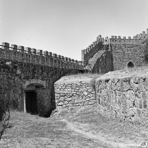 A black and white photograph depicts weathered stone town walls with arched entrances and stairways, set against a clear sky. The scene conveys a sense of history and resilience, with the stone walls showing signs of age and erosion. The afternoon light highlights the textures of the stonework.