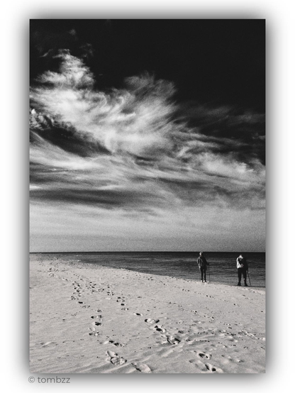 A black-and-white analogue photograph of the seashore beneath a vast sky filled with sweeping clouds. The composition unfolds in three layers: a spectacle of clouds above the horizon, a dark band of sea with two people at the water’s edge, and pale sand marked by footprints in the foreground.
The sky fills most of the frame. Bright clouds stretch diagonally, separating into delicate strands and weaving into broad, feathery streaks. Their soft edges dissolve into the deep, almost black expanse above. Visible grain enhances the image’s texture, while the rich tonal range, from luminous white through subtle greys to dense black, gives the sky remarkable depth.
On the right, two small figures with walking poles stand facing the sea. The calm water and straight horizon balance the movement of the clouds. Footprints lead the eye deeper into the photograph, towards the people and the open space.
There is both stillness and grandeur in this view. The two people seem tiny beneath the immense sky, yet their presence lends the landscape an intimate quality. An ordinary moment by the sea takes on the quiet power of an almost solemn encounter with nature.
