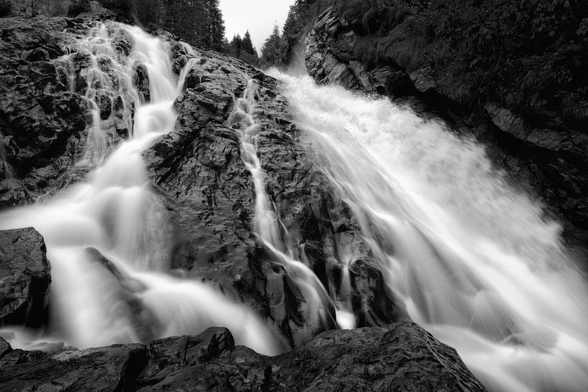 Schwarz-Weiss-Aufnahme eines kaskadierenden Wasserfalls in dramatischer Nahansicht. Das Wasser stürzt über dunkle, schroffe Felsformationen in die Tiefe und teilt sich dabei in mehrere sprudelnde Arme. Durch eine Langzeitbelichtung erscheint das fliessende Wasser weich, fast wie weisser Schleier oder Nebel, was einen starken Kontrast zu den nassen, dunklen Felsen bildet. Am oberen Bildrand sind schemenhaft Nadelbäume und Felswände zu erkennen.