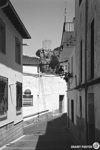 Narrow street between whitewashed buildings, leading to a distant castle on a hill. The scene is bright under a clear sky, creating a contrast between light and shadow on the cobblestone path.