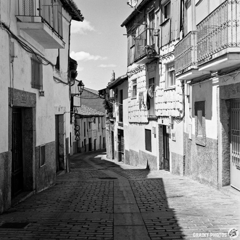 A black-and-white photograph depicts a narrow, cobblestone street ascending between tall buildings in Béjar, Spain. Afternoon light casts long shadows along the pavement, highlighting the texture of the stone buildings and their balconies. The scene evokes a sense of quiet history and timelessness.