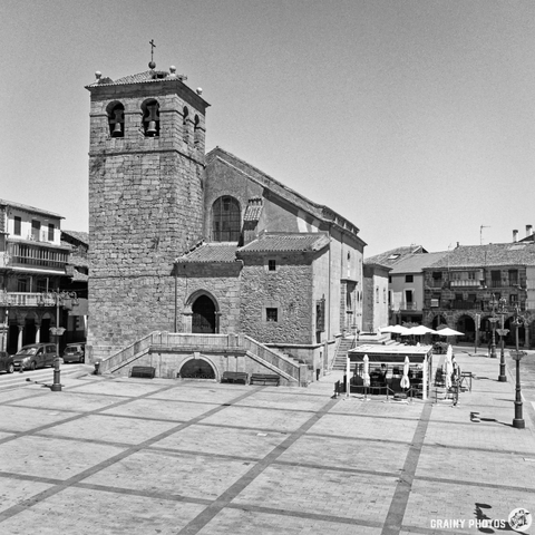 A black-and-white photograph shows the imposing stone church in Béjar, Spain, set against a clear blue sky. People are seated at outdoor cafes on the paved square in front of the church, and a few pedestrians stroll past. The afternoon light casts strong shadows across the square.