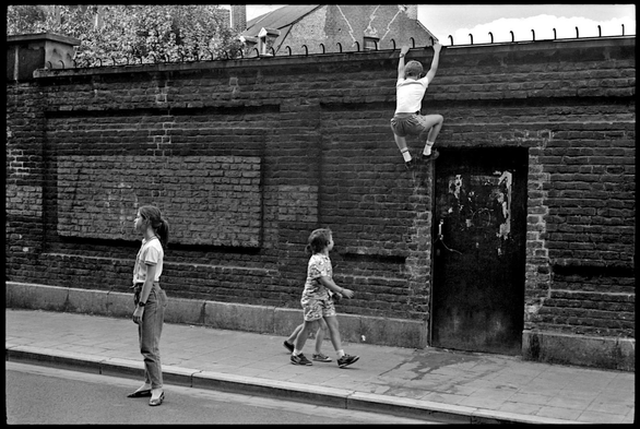 Scène de rue en n&b: un enfant escalade un mur de briques sombres pendant qu'un autre semble faire le guet pendant que passent 2 autres / Black-and-white street scene: a child is climbing a dark brick wall whilst another appears to be keeping watch as two others walk past.
