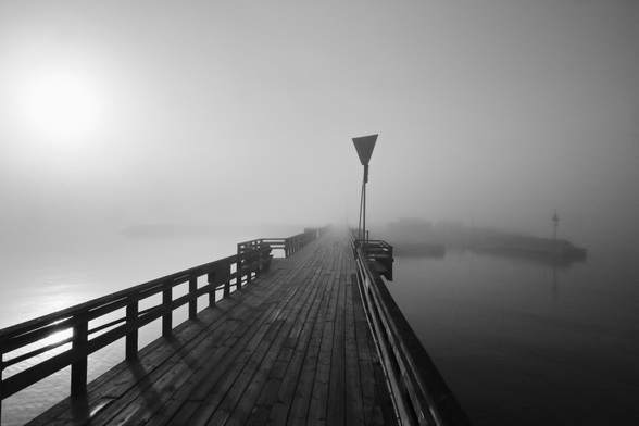 Schwarzweiss-Foto eines Holzsteges in die Ostsee,dessen Ende im Frühnebel und Gegenlicht der dahinter aufgehenden Sonne verschwimmt.