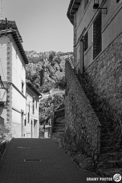 A narrow stone street lined with old buildings, leading up to a staircase. The background features rocky hills under a clear sky, creating a serene atmosphere in a historic setting. Black and white photography enhances the timeless charm of the scene.