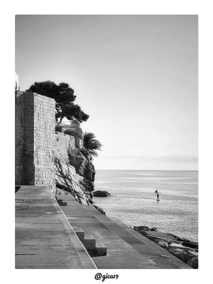 Es una fotografía vertical en blanco y negro de la muralla de Peñíscola sobre el mar. Una persona diminuta practica paddle surf en el agua tranquila.
La imagen se divide en dos mitades. A la izquierda dominan la piedra y la arquitectura, con muros, roca y vegetación. A la derecha hay un mar liso y un cielo muy despejado. Ese contraste entre masa y vacío da a la foto una sensación de calma y espacio.
La muralla es un bastión de sillería, con los sillares bien marcados por la luz lateral de la mañana. Ocupa el lado izquierdo y aporta peso visual. La muralla se apoya sobre una roca estratificada y agrietada, que cae hacia el mar con texturas muy definidas. Un pino mediterráneo de copa oscura y una palmera asoman por encima del muro. Sus siluetas destacan contra el cielo claro. Detrás de los árboles se ve una pequeña garita o torreta de vigilancia con cúpula, de estilo defensivo. Y en primer plano hay losas de hormigón que descienden en diagonal hacia el mar, con escalones. Esas líneas llevan la vista hacia el horizonte. A la derecha se amontonan bloques de roca del rompeolas.
En el mar, un poco a la derecha del centro, una figura minúscula rema de pie sobre su tabla. Es el único punto de interés humano y da escala a la escena. Cerca hay otra forma pequeña que es una embarcación.
La luz baja de la mañana crea un buen contraste. Los muros iluminados se ven claros y las sombras de los árboles y del espigón son profundas. El mar refleja un brillo suave y el horizonte queda alto y limpio, con una ligera franja de bruma. El blanco y negro resalta las texturas de la piedra y las líneas geométricas del espigón. El resultado es tranquilo y minimalista, con el contraste entre el patrimonio defensivo y el ocio moderno en el mar.
