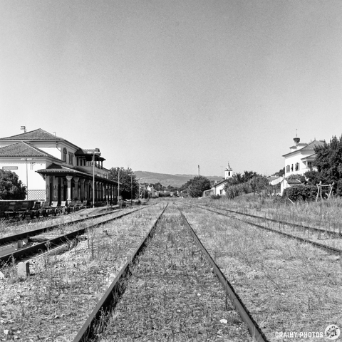 A black and white photograph depicts a long, disused railway line stretching towards two historic buildings under a bright midday sun. The tracks are overgrown with vegetation, leading the eye to a building with a covered platform and another further down the line, both characteristic of older European architecture. The scene evokes a sense of stillness and forgotten history.