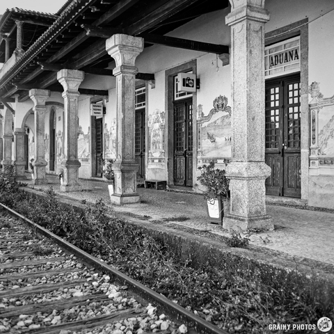 A black and white photograph depicts the facade of a historic customs house in the Beirã Railway Station, Portugal. The building features ornate stone pillars, decorative panels, and a tiled roof, with a railway track running along the front. The scene is bathed in bright midday light.