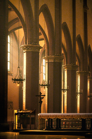 The photo if of an atmospheric interior La Basilica di San Francesco. It has  tall stone pillars and vaulted pointed arches illuminated by warm, golden light filtering through tall windows. In the foreground there is an altar draped in a patterned white cloth stands on a raised platform. Beside it on the left there is a lectern and a crucifix. Above in between the arches there are iron chandeliers which hang down between the large columns.