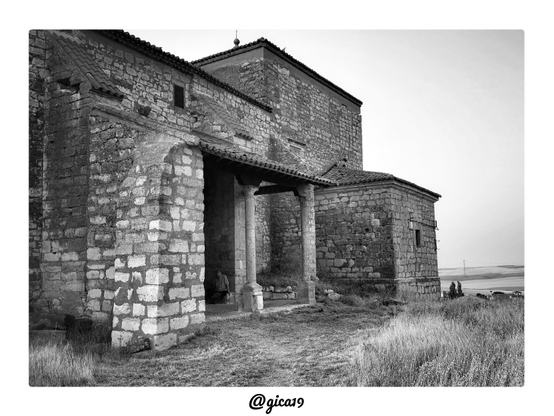 Esta foto la hice la tarde del 12 de agosto de 2026, justo antes del eclipse total de sol, en la Iglesia de San Esteban de Fuentesecas, en Zamora. Es una imagen en blanco y negro que muestra el lateral del templo, con sus muros de mampostería y un pórtico de entrada que sobresale de la fachada.
En primer plano, a la izquierda, se levanta un contrafuerte o machón de sillares irregulares. Los bloques más claros reciben la luz y destacan sobre el resto de la piedra, que está más oscura y desgastada. Desde ese volumen arranca un tejado de teja árabe en pendiente que cubre el pórtico. Bajo él se ven dos columnas de fuste liso con capiteles sencillos, apoyadas sobre basas de piedra. En la sombra del pórtico, junto a las columnas, hay una persona sentada que se distingue con dificultad.
Detrás del pórtico se alza el cuerpo principal de la iglesia, con muros de piedra de aspecto antiguo y una pequeña ventana estrecha. Más arriba aparece un volumen más alto, rematado por un tejado a cuatro aguas con una bola y una cruz en el vértice. A la derecha hay una dependencia más baja, también de piedra y con cubierta de teja, que tiene una ventana pequeña con reja y algunos elementos metálicos adosados al muro.
El templo está rodeado de hierba seca y alta, con un camino de tierra y hierba pisada que lleva hasta la entrada. A la derecha, al fondo, se abre el paisaje de campos de cereal ondulados, con unos pocos árboles y casas en la lejanía y una torre eléctrica en el horizonte. El cielo está despejado y con luz difusa, y la luz de la tarde resalta la textura de la piedra y de la hierba.
