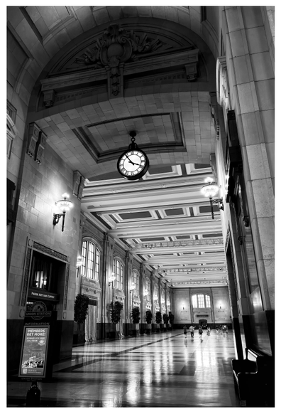 Black and white photo of the interior of Union Station, Kansas City. Tiny, distant figures walk along the wide corridor of a stone hall with soaring paneled ceilings. Wall-mounted lamps, and sunlight streaming through arched windows, are reflected on the polished floor. The scene is framed by a huge archway in the foreground, topped by an elaborate carved lunette. Below this is suspended a giant round clock with Roman numerals, its hands showing 3:55. Underneath the arch is a freestanding sign that says “Union Station” with information on membership.