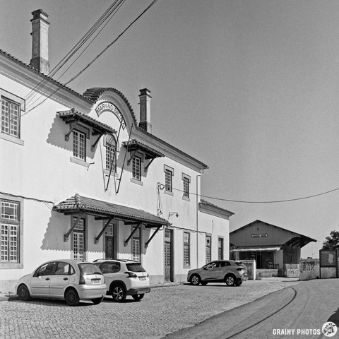 A black and white photograph shows a grand, two-story train station building with intricate architectural details. Several cars are parked in front of the station, and a paved road curves away into the distance under bright midday sunlight. The scene evokes a sense of timelessness and quiet observation.