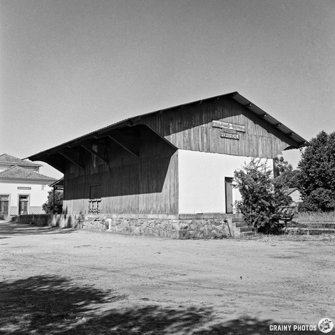 A black and white photograph depicts a large, weathered wooden train station building in Beirã, Portugal. The structure features a covered platform and signage indicating its location, set against a backdrop of trees and a paved area. The morning light illuminates the building's facade, creating a sense of quiet history and rural charm.