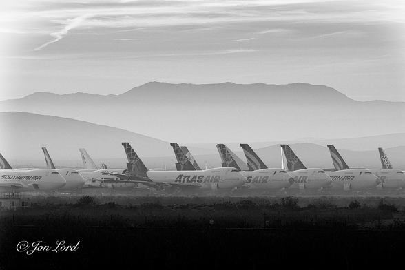This is a black and white dawn photo in landscape format of a large number of parked Boeing 747 aircraft arranged in rows in longterm storage with a mountain range in the background. Mojave Air and Space Port, California (2016).
In the foreground and rising up to a little less than the quarter way up level is desert brush mostly deep in shadow. Above are the neat rows and lines of those parked Boeing 747 aircraft. They are view obliquely with the noses and right side in view and observed from a few hundred metres away. All the planes have their noses pointing to the right. The closest aircraft have a white fuselage and dark tail with the words 'Atlas Air' in large lettering in the centre of the fuselage. In the row behind, the aircraft again have similar colouring but with the words 'Southern Air' in large dark lettering. Most have their forward cargo doors open. Many have engines and other parts removed. In the background the air is hazy with a distant tall and long mountain range stretching across the photo from left to right. Above is a partly cloudy sky with only thin wisps of cirrus high clouds obscuring the heavens.