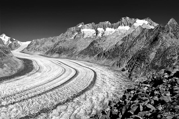This monochrome photograph captures the vast expanse of the Aletsch Glacier winding through a rugged mountain range. Deep longitudinal ridges are carved into the frozen surface, tracing the glacier's path towards the valley floor. In the foreground, a field of coarse rocks provides a contrasting texture to the glacier's smooth flow. Beyond the snow, craggy peaks and snow-dusted summits rise sharply into the sky. The interplay of light and shadow defines every fissure and ridge within the frozen terrain. This scene captures the enduring scale of a landscape shaped by time.