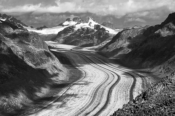 A wide glacier flows through a mountain pass, its surface marked by prominent longitudinal ridges that curve toward the lower right of the frame. These icy paths are flanked by steep, craggy slopes composed of jagged rock and fragmented debris. The rough texture of the stone contrasts with the repetitive, linear patterns carved into the glacial bed. In the distance, snow-capped peaks rise against a heavy sky filled with textured clouds. A monochromatic palette defines the scene, highlighting the stark interplay of light and shadow across the rugged landscape.