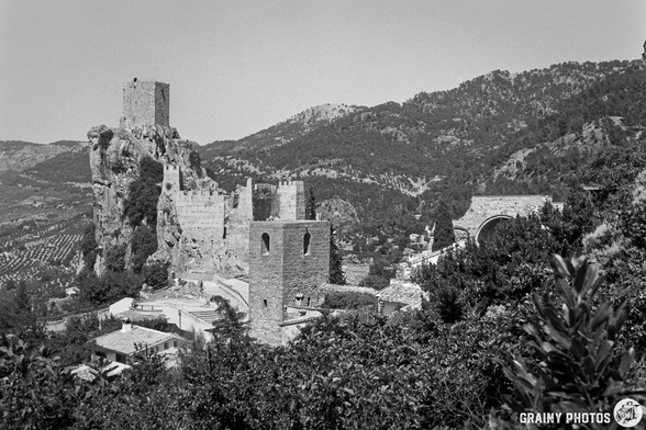 A black and white photograph of La Iruela castle ruins perched on a rocky hill, surrounded by lush greenery and mountainous terrain in the background. The structure features tall stone towers and remnants of walls, evoking a sense of historical significance.