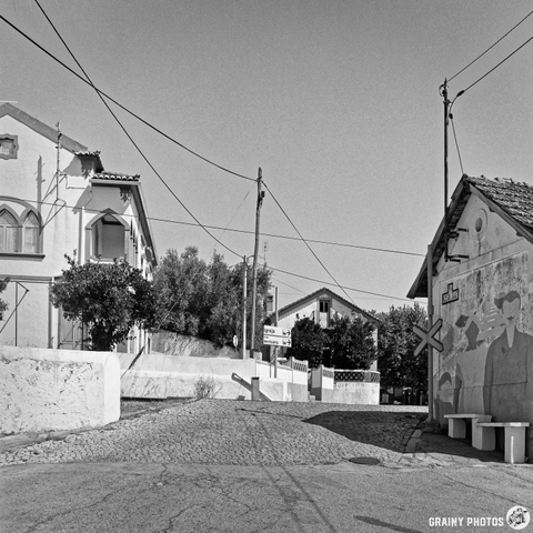 A black and white photograph captures a tranquil street and level crossing in Beirã, Alentejo, Portugal. The scene features traditional white buildings with tiled roofs, framed by overhead power lines and a small grassy area with signage. The midday sun casts strong shadows, creating a sense of stillness and timelessness.