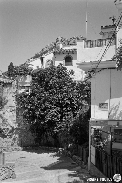 A scenic narrow street in La Iruela featuring charming white buildings, lush greenery, and a rocky hillside in the background. The image is in black and white, adding a timeless feel to the serene atmosphere.