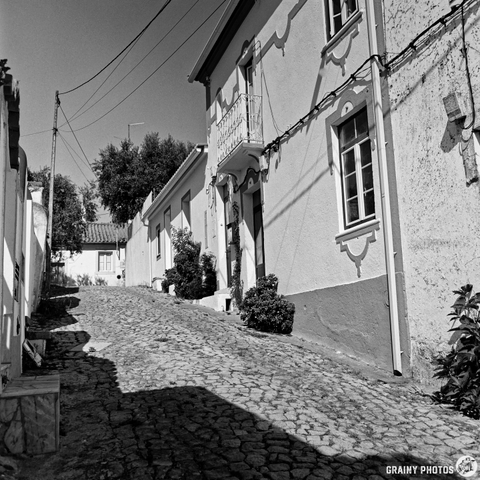 A black and white photograph depicts a narrow, cobbled street lined with traditional white houses in Beirã, Portugal. The late morning sunlight casts long shadows across the street, highlighting the texture of the cobblestones and the architectural details of the buildings. The scene evokes a sense of quiet charm and timelessness.