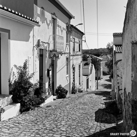 A narrow, cobblestone street slopes downwards between weathered buildings in BeirãAlentejo, Portugal. The black and white photograph captures the textures of the stone walls, the uneven pavement, and a small patch of greenery at the base of one building. The midday sun casts strong shadows along the street, creating a sense of depth and quietude.