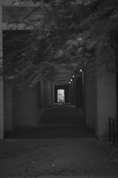 Photo looking down an outdoor corridor, a bright rectangle of sunlight visible at the far end.