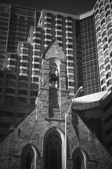 Photo of a stone church nestled in amongst taller, modern buildings.
