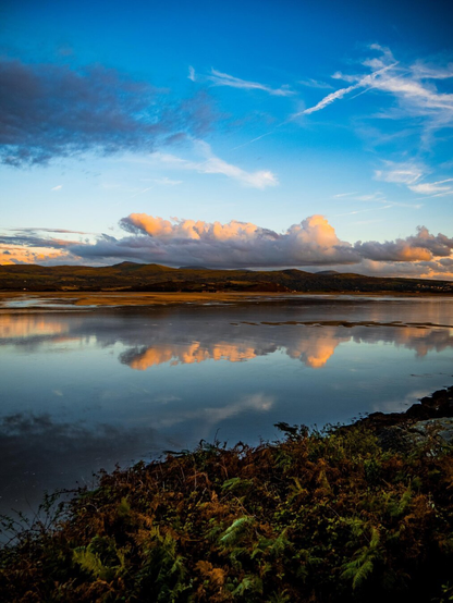 A landscape shot of calm waters off the coast of North Wales at dusk. Vibrant orange and white clouds are reflected in the still blue water. In the foreground, a rocky, autumnal fern-covered bank is lit by golden hour sunlight and in the distance mountains rise under the moody, low hanging clouds.