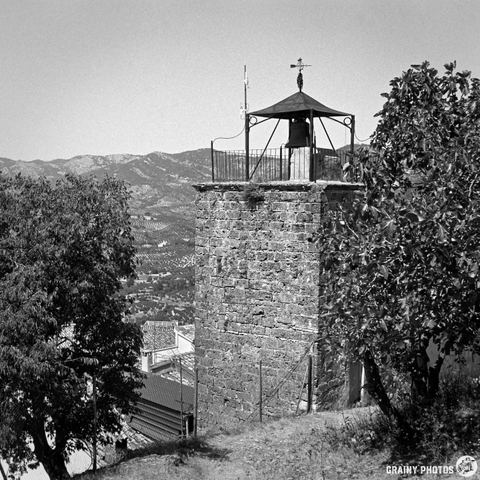 A stone tower with a bell and a cross, surrounded by trees, perched on a hillside with distant mountains in the background. The image is in black and white, highlighting the architectural details and natural scenery.