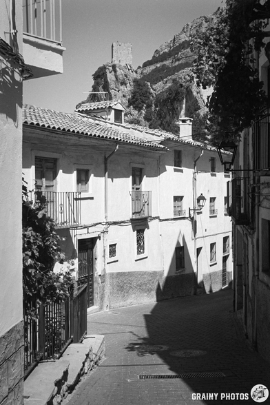 A narrow cobblestone street lined with whitewashed buildings, featuring wrought iron balconies. In the background, a castle ruins stands atop a rocky hill, under a clear sky. The scene captures a charming historic town atmosphere in black and white.