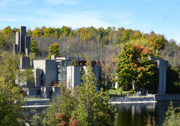 Modernist concrete buildings surrounded by lots of trees are seen along the shoreline of a river. The tops of the trees show the orange and red of the changing season on this bright and sunny day.