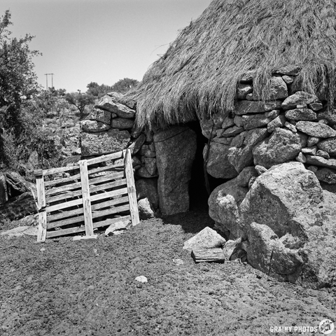 A black-and-white photograph shows a weathered stone structure with a thatched roof, possibly a shepherd’s hut. A simple wooden gate stands to the side, leading into a dark opening in the stone walls, set against rolling hills and sparse vegetation. The afternoon light casts long shadows across the ground, creating a sense of timelessness and solitude.