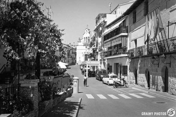 A quiet street scene in a charming town, featuring narrow pathways, balconies with plants, a church in the background, and a pedestrian crossing, all captured in black and white.