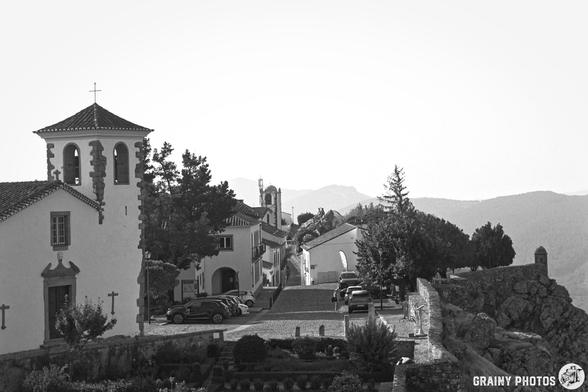 A black and white photograph shows a charming, historic village perched on a hilltop. Traditional buildings with tiled roofs cluster around a prominent tower, overlooking rolling hills and distant mountains under bright morning light. A stone wall runs along the foreground, adding to the picturesque scene.