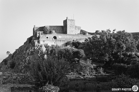 A black-and-white photograph shows a stone castle perched atop a rocky hill, surrounded by lush greenery. The castle's walls and towers are visible against a bright sky, creating a strong contrast of light and shadow. The scene evokes a sense of history and timelessness.