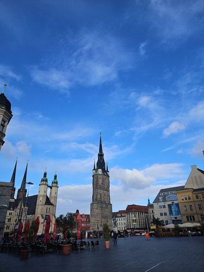 Hochkant Foto eines leeren Marktplatzes. Zusammengefaltete Sonnenschirme neben leeren Tischen und Stühlen rahmen schwarze Steinplatten ein. Zentral im Hintergrund steht ein einzelner Turm mit Uhr und ragt in den tiefblauen Himmel. Links daneben ein Dom mit weiteren Türmen. Ein paar einzelne dünne Wölkchen werden von der schwachen Morgensonne erleuchtet. Es sieht kühl aus. 