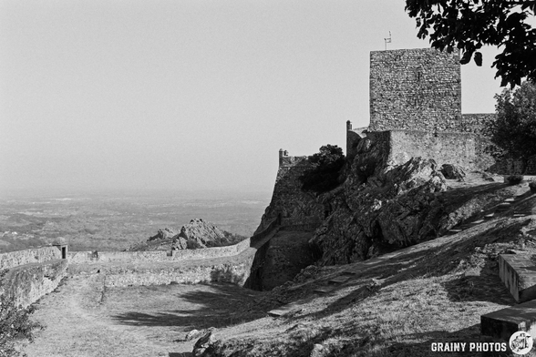 A black-and-white photograph shows a stone castle and the keep perched atop a rocky outcrop overlooking rolling hills and a distant landscape. The scene is bathed in bright morning light, highlighting the textures of the stone walls and the rugged terrain. A winding path along the castle wall leads along the cliff edge, adding depth to the composition.