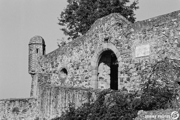 A black-and-white photograph shows a stone archway and tower, part of an old fortification overlooking the landscape. The stonework is detailed with a rough texture and visible vegetation growing along the walls, captured on a clear morning.