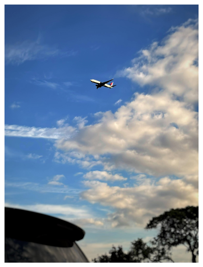 Photo taken from a car window, of a distant commercial airliner descending through a blue sky that’s partly covered with clouds. A contrail streak from a different plane stretches from a cloud towards left frame. In the foreground are blurred trees and the back window of a car in the neighboring lane.