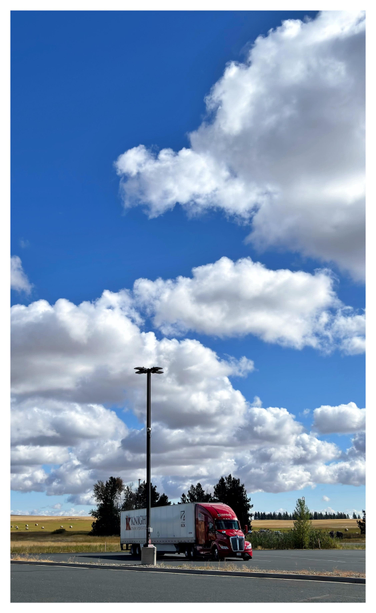 Portrait-orientation photo of a trailer truck with a red cab pulling a long white cargo box that says “Knight Transportation,” parked in an empty lot beside a tall streetlight in front of a flat Idaho landscape. Above, herds of cumulus clouds cross a wide blue sky.