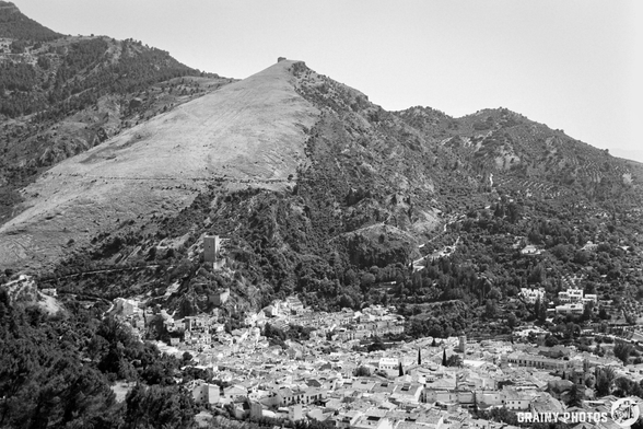 A panoramic view of a mountainous landscape featuring Cazorla nestled below. The terrain is predominantly rocky with sparse vegetation, showcasing a blend of natural beauty and human settlement under a clear sky in black and white tones.