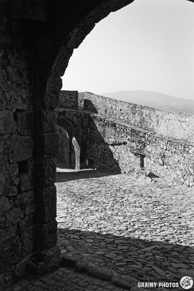 A black-and-white photograph shows a stone archway framing a courtyard with cobblestone paving. Beyond the arch, a low wall and distant hills are visible under bright morning light, creating strong shadows across the foreground. The scene evokes a sense of history and tranquillity.